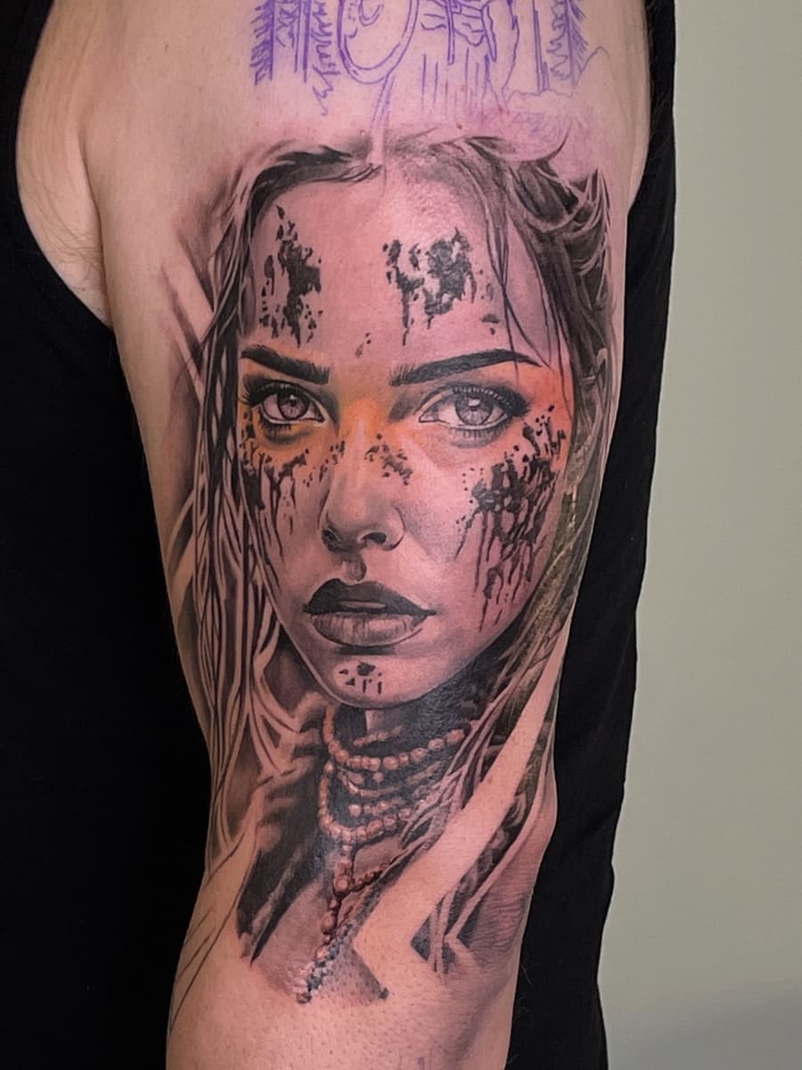 Black-and-grey portrait tattoo on upper arm showing a woman's face with dark paint-like splashes across the forehead and cheeks, detailed eyes, layered necklaces, and soft shading.