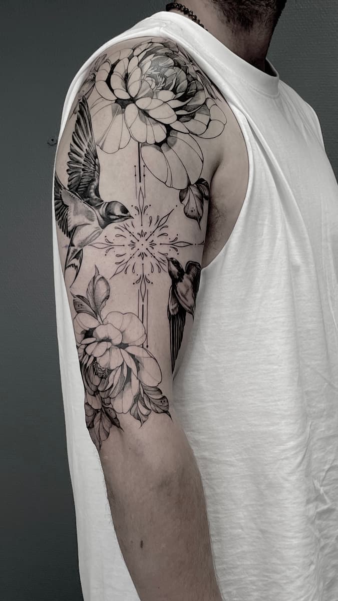 Black-and-grey upper arm tattoo with large flowers, two birds in flight, and a central ornamental geometric design with fine line details.