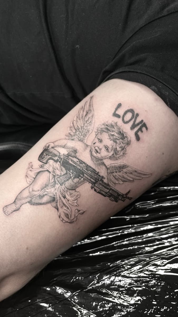 Black-and-grey cherub tattoo holding a rifle on the upper arm, with the word "LOVE" above in bold handwritten-style lettering.