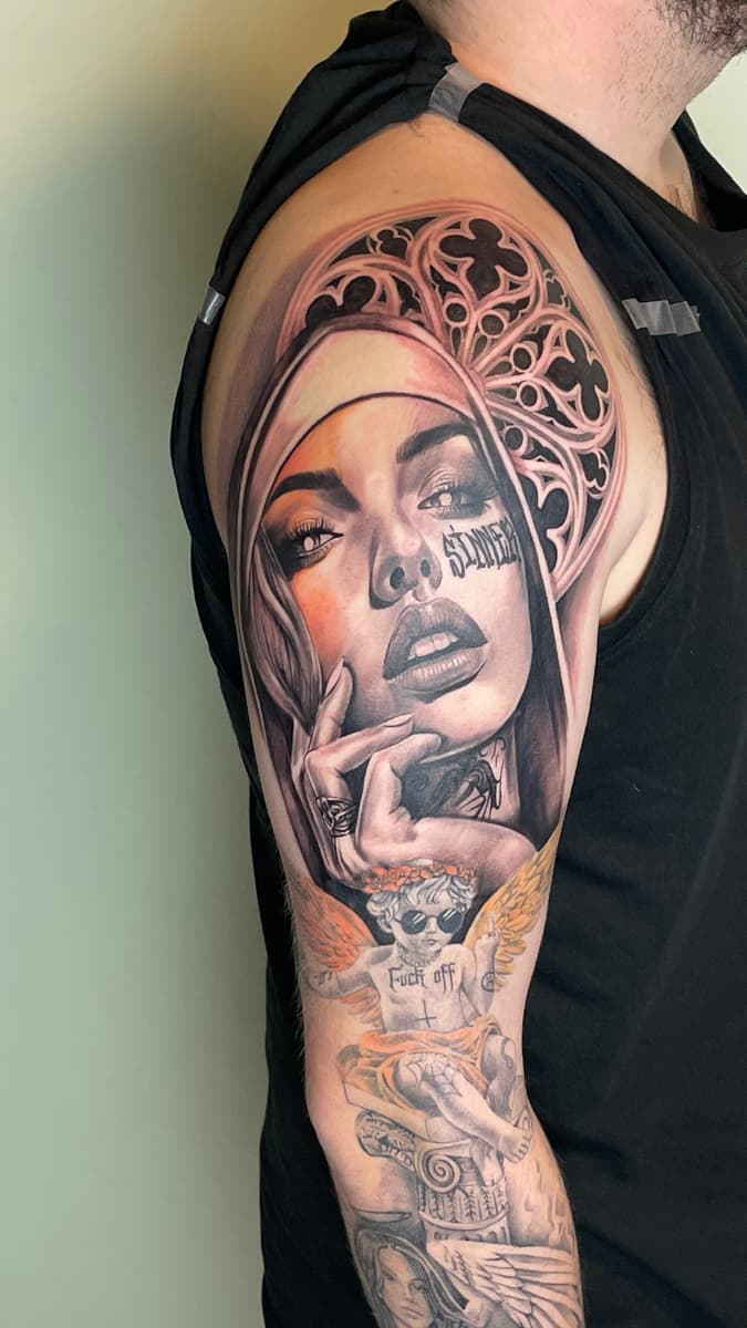 Large upper arm portrait tattoo of a woman in black-and-grey with warm orange highlights, framed by gothic-style ornamentation, above an existing cherub tattoo.
